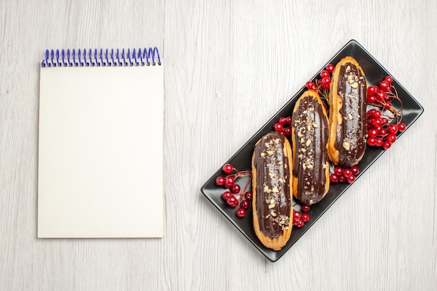 Vue de dessus des éclairs au chocolat et des raisins de Corinthe sur la plaque rectangulaire isométrique noire sur le côté droit et un cahier sur le côté gauche du sol en bois blanc