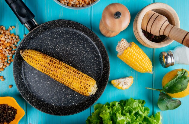 Vue de dessus d'épis de maïs dans une casserole avec des graines de maïs graines de poivre noir couper le sel de maïs citron et laitue autour de la surface bleue