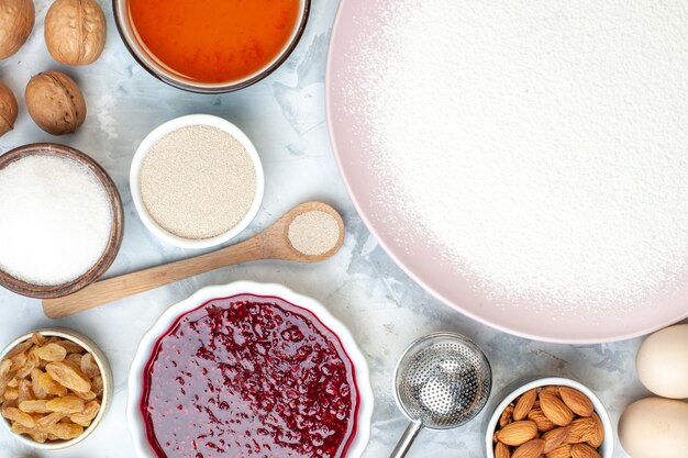 Vue de dessus farine en poudre sur assiette bols avec graines de sésame amandes confiture oeufs cuillère en bois sur table