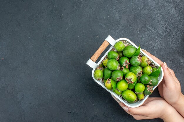 Vue de dessus feijoas frais sur panier en plastique dans la main féminine sur une surface sombre avec copie espace