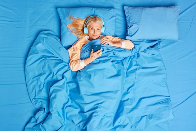 Vue de dessus de la femme d'âge moyen utilise un smartphone et des écouteurs stéréo pour écouter de la musique relaxante pose dans un lit confortable bénéficie d'un nouveau jour a assez de sommeil se trouve sur un oreiller doux sous une couverture bleue