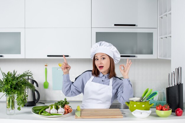 Vue de dessus d'une femme chef confiante et de légumes frais pointant vers le haut en faisant un geste de lunettes dans la cuisine blanche