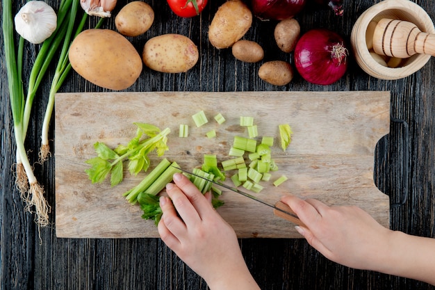 Vue de dessus de femme main couper le céleri sur une planche à découper avec des légumes et un broyeur d'ail sur fond de bois