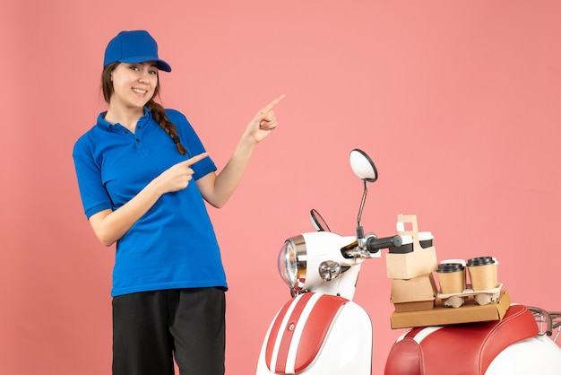 Vue de dessus d'une femme de messagerie souriante debout à côté d'une moto avec du café et de petits gâteaux pointant vers le haut sur la couleur pêche pastel