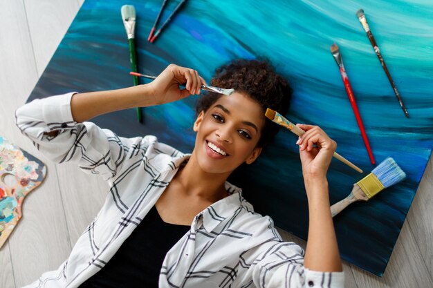 Vue de dessus d'une femme peintre heureuse couchée sur une toile avec des pinceaux dans les mains. Rêver et se détendre après un travail productif.