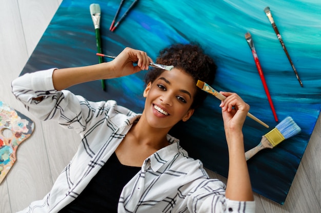 Vue de dessus d'une femme peintre heureuse couchée sur une toile avec des pinceaux dans les mains. Rêver et se détendre après un travail productif.
