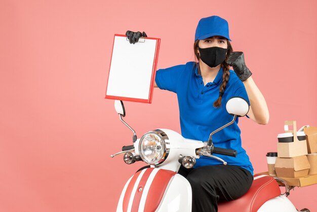 Vue de dessus d'une fille de messagerie confuse portant un masque médical et des gants assis sur un scooter tenant une feuille de papier vide livrant des commandes sur fond de pêche pastel