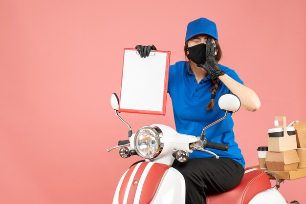 Vue de dessus d'une fille de messagerie émotionnelle portant un masque médical et des gants assis sur un scooter tenant une feuille de papier vide livrant des commandes sur fond de pêche pastel