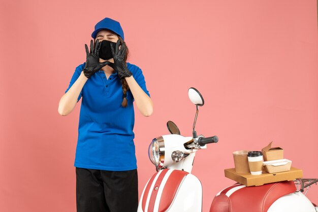 Vue de dessus d'une fille de messagerie portant des gants de masque médical debout à côté d'une moto avec du café dessus un gâteau appelant quelqu'un sur un fond de couleur pêche pastel