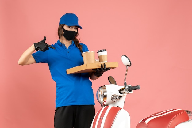 Vue De Dessus D'une Fille De Messagerie Portant Des Gants De Masque Médical Debout à Côté D'une Moto Pointant Du Café De Petits Gâteaux Sur Fond De Couleur Pêche Pastel