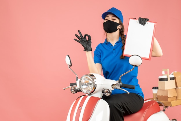 Vue de dessus d'une fille de messagerie rêveuse portant un masque médical et des gants assis sur un scooter tenant une feuille de papier vide livrant des commandes sur fond de pêche pastel