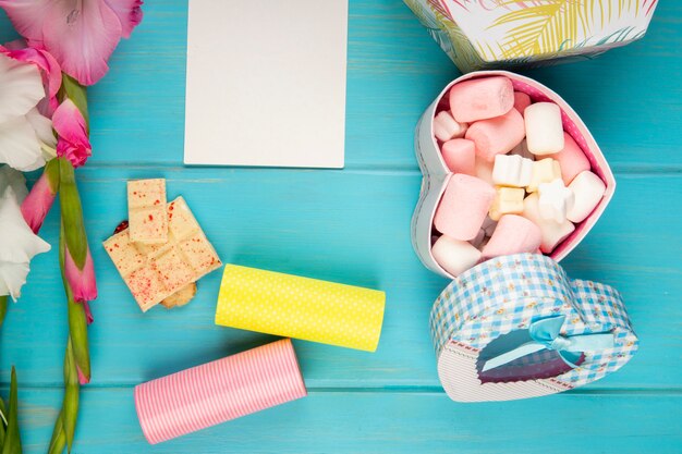 Vue de dessus de fleur de glaïeul de couleur rose avec rouleau de ruban adhésif, feuille de papier blanc, barre de chocolat blanc et boîte-cadeau colorée remplie de guimauve sur table bleue
