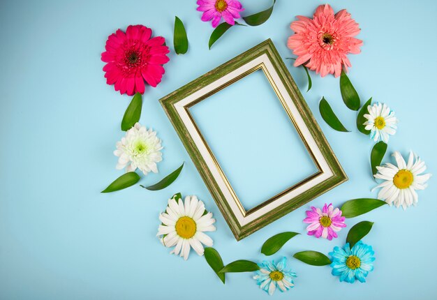 Vue de dessus des fleurs colorées de gerbera avec marguerite et feuilles de ruscus disposées autour d'un cadre vide sur fond bleu avec copie espace