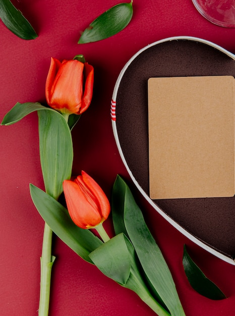 Vue de dessus des fleurs de tulipe de couleur rouge avec boîte-cadeau en forme de coeur avec une carte postale ouverte sur fond rouge