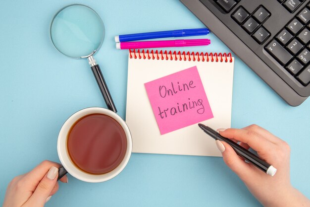 Vue de dessus formation en ligne écrite sur pense-bête tasse de thé et marqueur dans les mains des femmes clavier lupa bloc-notes marqueurs bleus et roses sur bleu