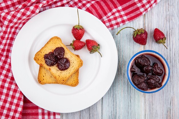 Vue de dessus de fraise sur une plaque blanche sur un tissu à carreaux rouge avec une confiture de fraises sur un bol sur un fond de bois gris