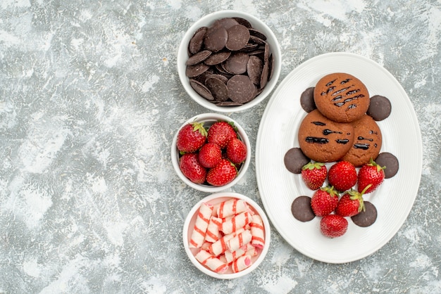 Vue de dessus, fraises, biscuits au chocolat et chocolats ronds sur la plaque ovale blanche et bols avec des bonbons, des fraises et des chocolats sur le côté droit du sol