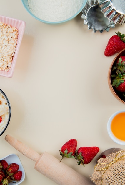 Vue de dessus des fraises dans un bol et du beurre fondu avec de la farine et de l'avoine avec un rouleau à pâtisserie sur une surface blanche avec copie espace