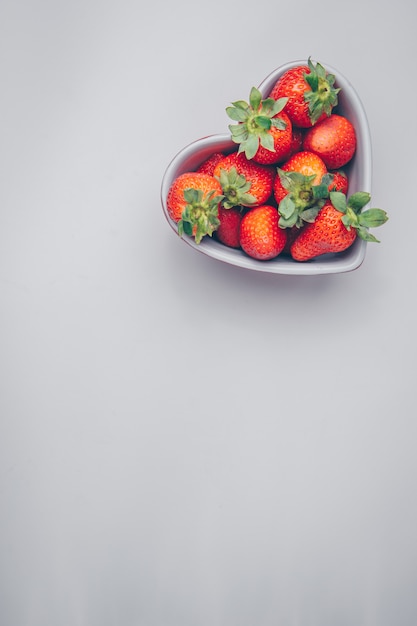 Vue de dessus des fraises dans un bol en forme de coeur sur fond blanc. espace libre vertical pour votre texte