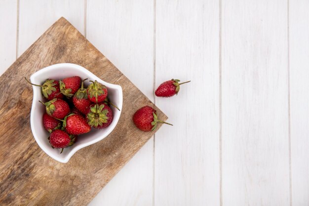 Vue de dessus de fraises fraîches sur un bol blanc sur une planche de cuisine en bois sur un fond en bois blanc avec espace copie