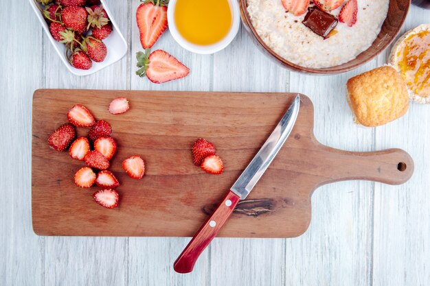 Vue de dessus de fraises mûres fraîches sur une planche de bois avec couteau de cuisine et bouillie d'avoine au miel dans un bol en bois sur rustique