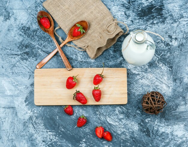 Vue de dessus fraises sur une planche à découper avec du lait, point d'écoute, cuillères en bois et un morceau de sac sur une surface en marbre bleu foncé. horizontal