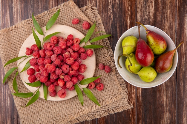 Vue de dessus des framboises avec des feuilles sur une planche à découper sur un sac et un bol de pêche sur une surface en bois