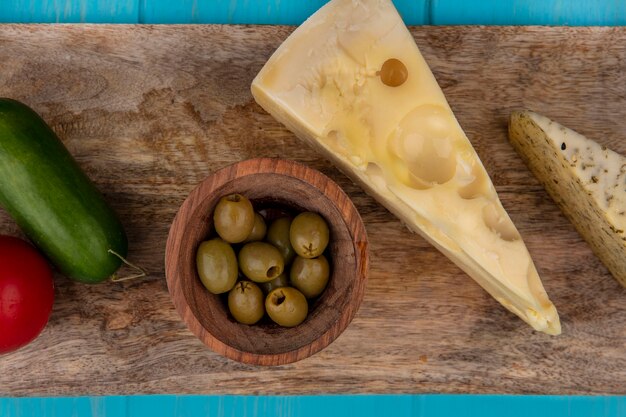 Vue de dessus fromage maasdam aux olives vertes dans des bols sur un support et concombre à la tomate