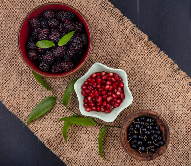 Vue de dessus des fruits comme les baies de grenade et de prunelle mûre dans des bols avec des feuilles sur un sac sur une surface noire