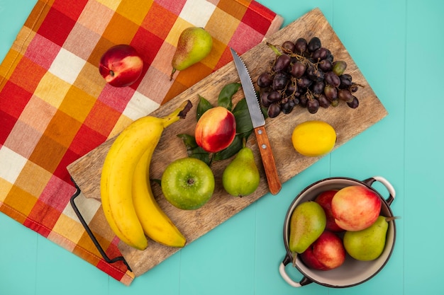 Vue de dessus des fruits comme banane pomme pêche poire citron raisin avec couteau et feuilles sur planche à découper sur tissu à carreaux avec pot de pêche poire sur fond bleu