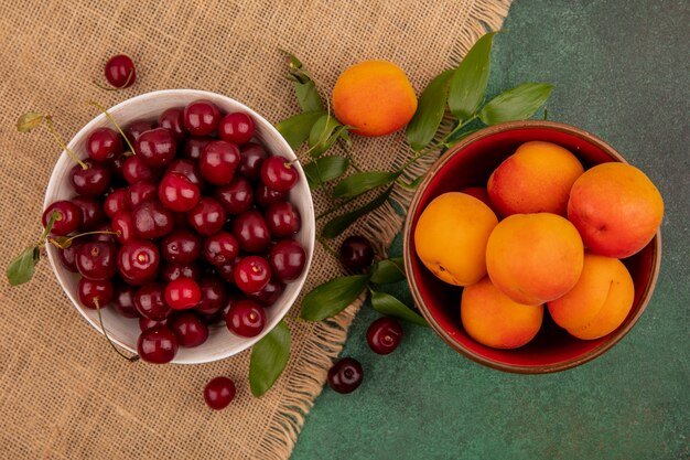 Vue de dessus des fruits comme les cerises et les abricots dans des bols avec des feuilles sur un sac sur fond vert