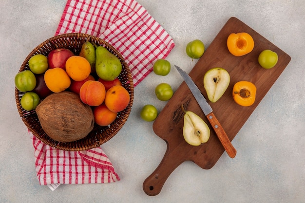 Vue de dessus des fruits comme la moitié de la poire et de l'abricot coupés avec un couteau sur une planche à découper et un panier de noix de coco pêche prune poire sur tissu à carreaux et prunes sur fond blanc