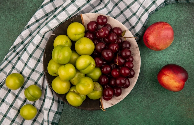 Vue de dessus des fruits comme les prunes et les cerises dans la plaque sur un tissu à carreaux avec des pêches sur fond vert