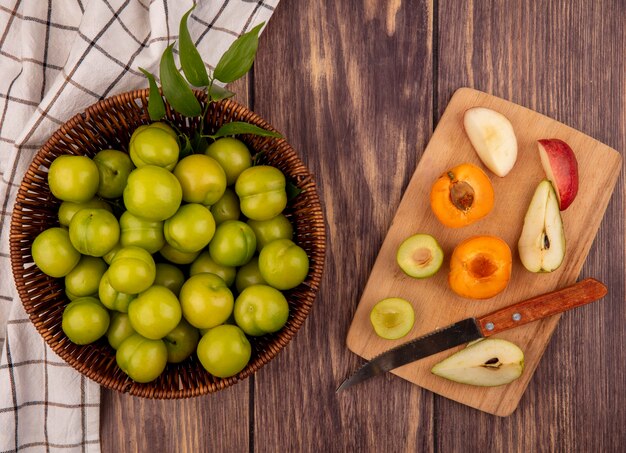 Vue de dessus des fruits comme des prunes vertes dans le panier sur un tissu à carreaux et demi-coupe abricot poire prune pêche avec un couteau sur une planche à découper sur fond de bois