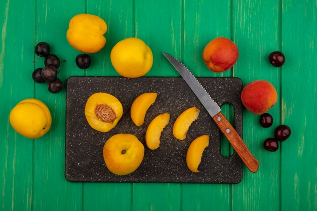 Vue de dessus des fruits entiers demi abricots tranchés avec un couteau sur une planche à découper et entiers avec des baies de prunelle sur fond vert