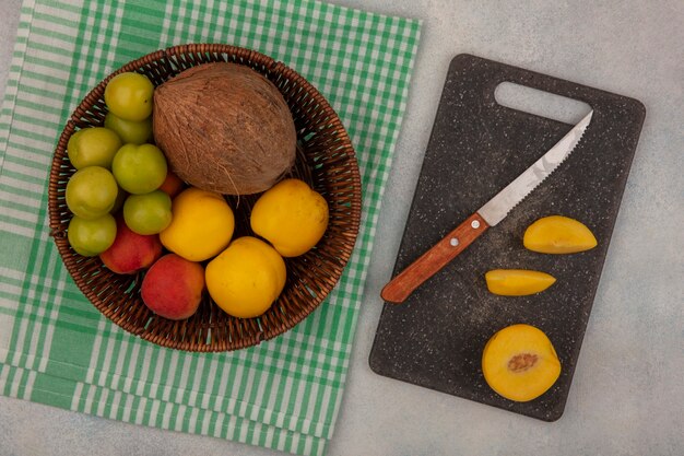 Vue de dessus de fruits frais tels que la cerise verte prune peachescoconut frais sur un seau avec des tranches de pêche sur une planche à découper de cuisine avec un couteau sur un fond blanc