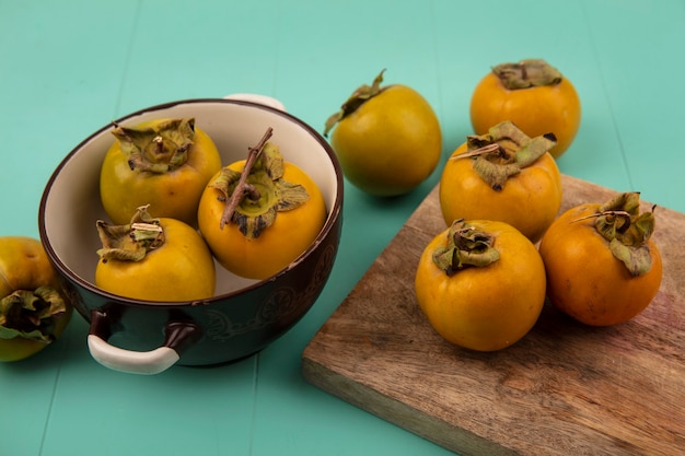 Vue de dessus des fruits de kaki arrondis orange sur une planche de cuisine en bois avec des fruits de kaki sur un bol sur une table en bois bleu