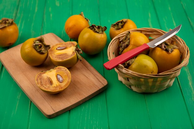 Vue de dessus des fruits de kaki sur un seau avec un couteau avec des fruits de kaki coupés en deux sur une planche de cuisine en bois sur une table en bois verte