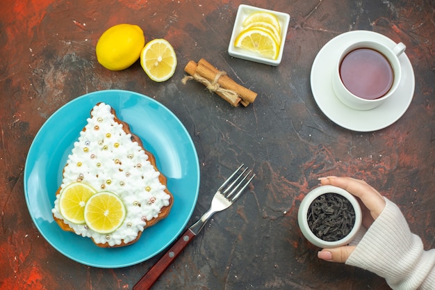 Vue de dessus gâteau au citron sur plaque bleue tasse de thé tranches de citron dans un bol fourchette à thé dans un bol dans des bâtons de cannelle main féminine sur fond rouge foncé