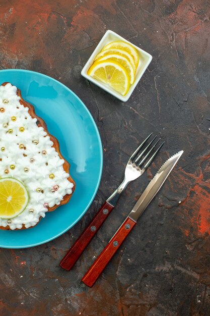 Vue de dessus gâteau au citron sur plaque bleue tranches de citron dans un bol couteau et fourchette sur fond rouge foncé