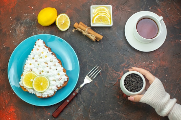 Vue de dessus gâteau au citron sur plaque bleue tranches de citron dans un bol fourchette tasse de thé bâtons de cannelle citron sur fond rouge foncé