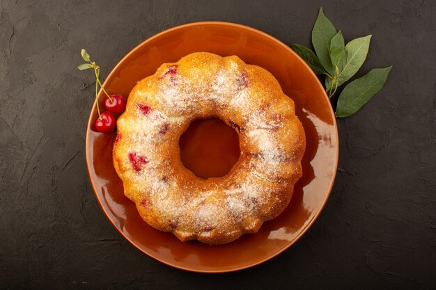 Une vue de dessus gâteau aux fruits cuit délicieux rond avec des cerises rouges à l'intérieur et du sucre en poudre à l'intérieur d'une plaque brune ronde