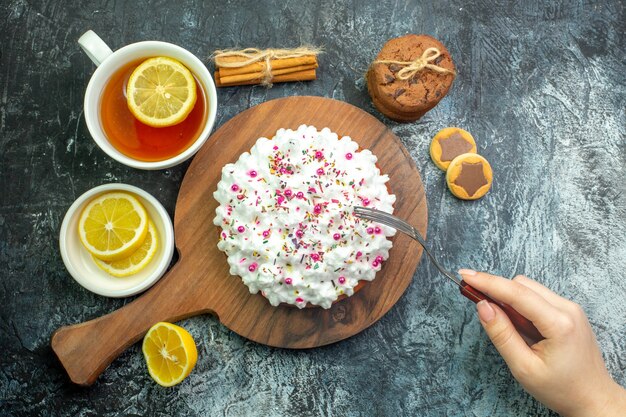 Vue de dessus gâteau avec crème pâtissière sur planche de bois biscuits bâtons de cannelle tasse de fourchette à thé dans la main féminine sur table grise