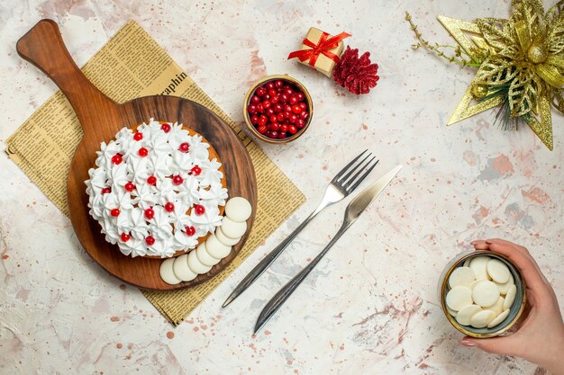 Vue de dessus gâteau avec crème pâtissière sur planche de bois sur papier journal ornement de Noël couteau et fourchette bol avec chocolat blanc dans la main féminine