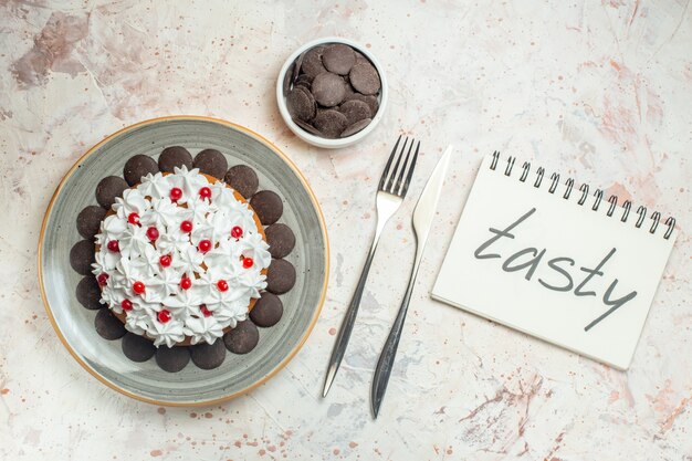 Vue de dessus gâteau avec crème pâtissière sur plaque ovale chocolat dans un bol fourchette et couteau à dîner. mot savoureux écrit sur ordinateur portable