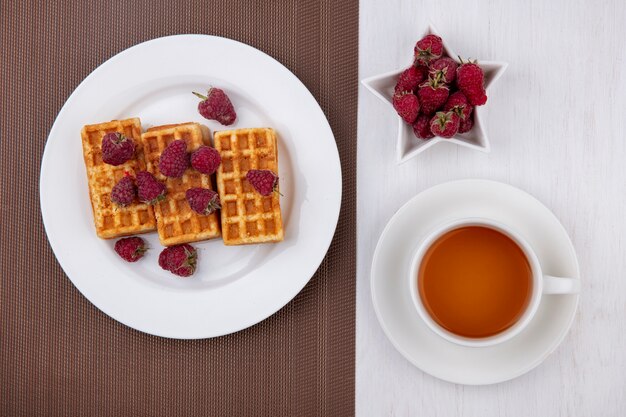 Vue de dessus des gaufres sur une assiette avec des framboises et une tasse de thé sur un tableau blanc