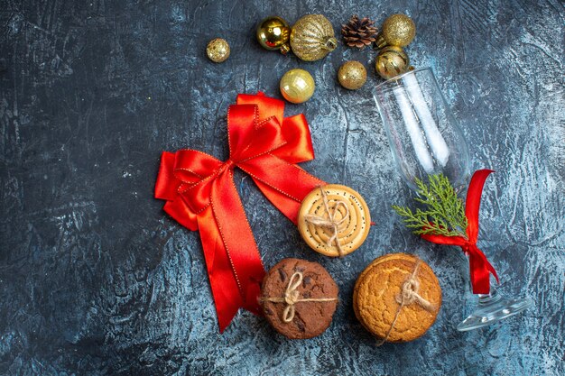 Vue de dessus d'un gobelet en verre tombé avec un ruban rouge et des accessoires de décoration à côté de biscuits empilés et d'un ruban en forme d'arc sur fond sombre