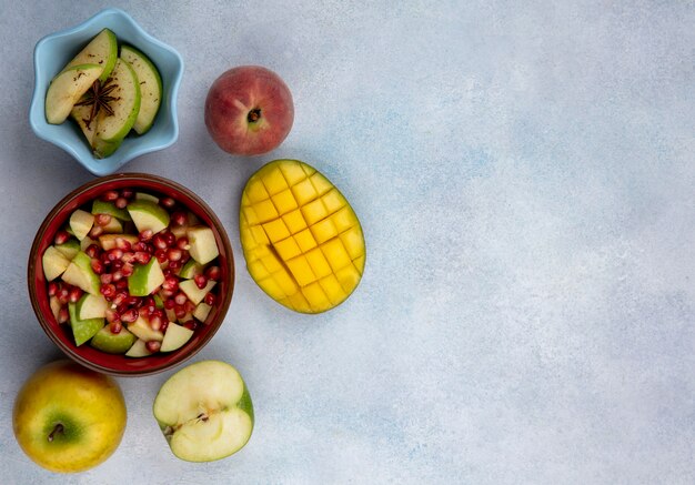 Vue de dessus des graines de grenade et des pommes hachées dans un bol rouge avec des tranches de mangue et de fruits frais sur une surface blanche