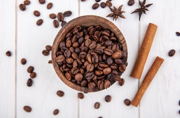 Vue de dessus des grains de café frais sur un bol en bois avec des bâtons de cannelle et d'anis sur un fond en bois blanc