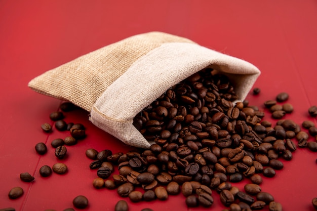 Vue de dessus des grains de café torréfiés tombant d'un sac de jute sur fond rouge